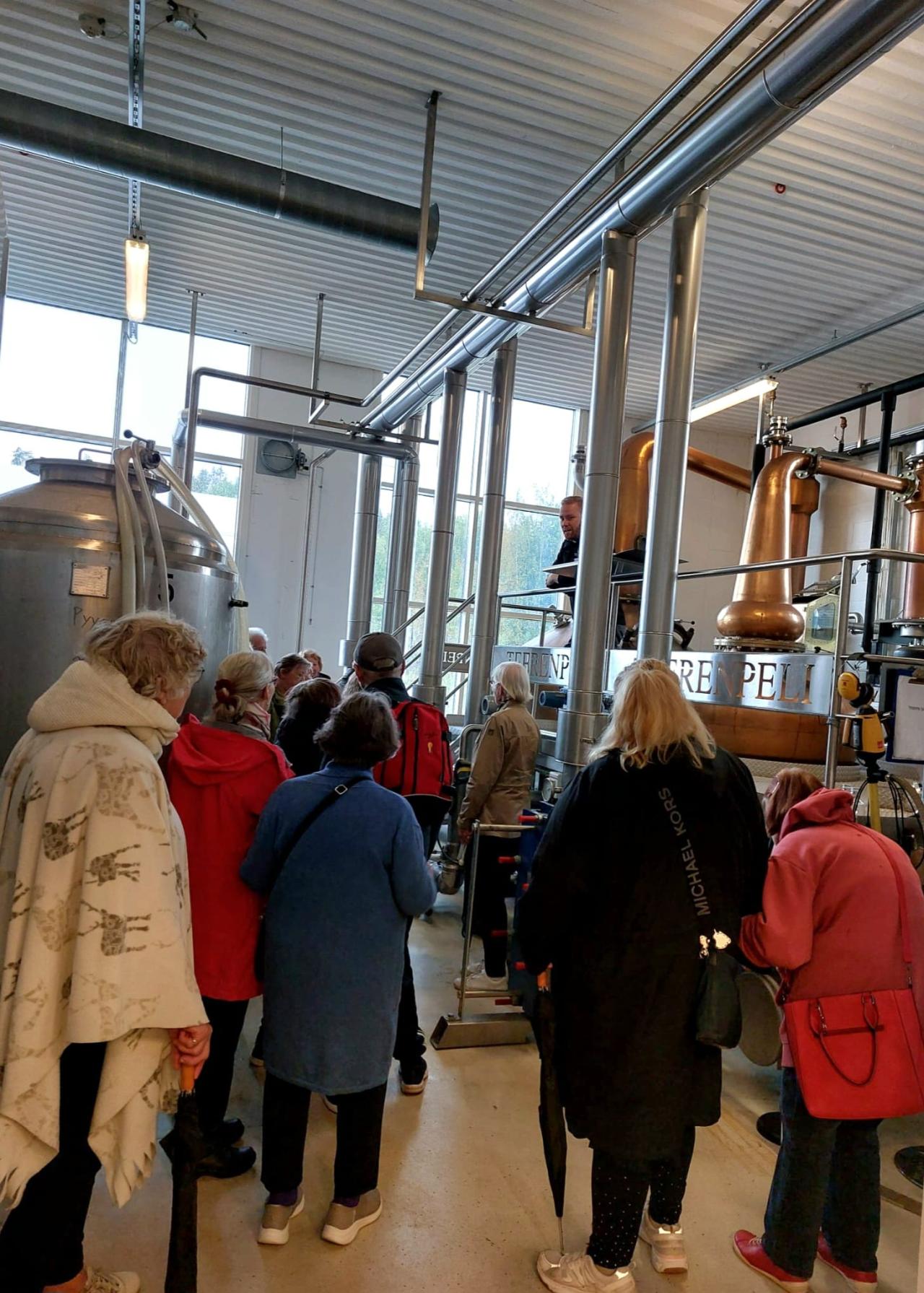 Kuva tuotantotiloista. Kuvassa metallisia putkia katonrajassa, josta laskeutuvat maahan. Keskellä kuvaa ihmisiä selät kameraan päin kuuntelemassa korokkeella seisovaa esittelijää.